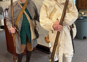 Full-length indoor shot of two older men posing in detailed historical attire from the American Revolutionary War period. The man on the left wears a brown tricorn hat, round spectacles, a long grey wool coat over a buttoned emerald green vest, tan breeches, brown stockings, and dark slip-on shoes. He grips a long wooden flintlock musket resting on the carpet. The man on the right wears a black tricorn hat with light trim, glasses, a mustache, a heavily fringed cream-colored linen hunting frock or rifle shirt with a matching sash cross-belt, white breeches, and tall black gaiters over his shoes. He cradles a similar long flintlock musket in his hands. They are standing on grey carpeting inside a public building or museum hall.