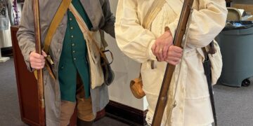 Full-length indoor shot of two older men posing in detailed historical attire from the American Revolutionary War period. The man on the left wears a brown tricorn hat, round spectacles, a long grey wool coat over a buttoned emerald green vest, tan breeches, brown stockings, and dark slip-on shoes. He grips a long wooden flintlock musket resting on the carpet. The man on the right wears a black tricorn hat with light trim, glasses, a mustache, a heavily fringed cream-colored linen hunting frock or rifle shirt with a matching sash cross-belt, white breeches, and tall black gaiters over his shoes. He cradles a similar long flintlock musket in his hands. They are standing on grey carpeting inside a public building or museum hall.