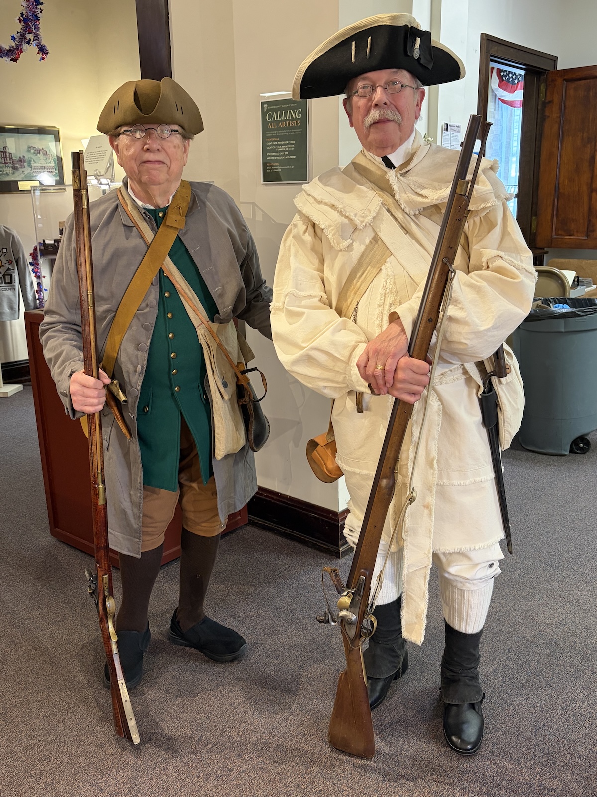 Full-length indoor shot of two older men posing in detailed historical attire from the American Revolutionary War period. The man on the left wears a brown tricorn hat, round spectacles, a long grey wool coat over a buttoned emerald green vest, tan breeches, brown stockings, and dark slip-on shoes. He grips a long wooden flintlock musket resting on the carpet. The man on the right wears a black tricorn hat with light trim, glasses, a mustache, a heavily fringed cream-colored linen hunting frock or rifle shirt with a matching sash cross-belt, white breeches, and tall black gaiters over his shoes. He cradles a similar long flintlock musket in his hands. They are standing on grey carpeting inside a public building or museum hall.