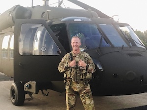 A full-length portrait of a smiling soldier standing on a tarmac directly in front of a large, dark green military helicopter. The soldier wears full operational camouflage pattern (OCP) uniform, tactical vest gear, and holds a radio or equipment attached to his chest vest. The helicopter's cockpit door on the left side is wide open, exposing the interior cabin. The shot is captured in soft, bright daylight with a clear background.