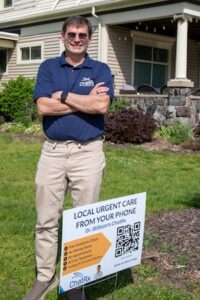 A smiling middle-aged man with glasses, a blue polo shirt, and beige khaki pants stands outdoors with his arms crossed on a grassy lawn. He is standing next to a white yard sign promoting "Local Urgent Care From Your Phone" and "Dr. Stillson's ChatRx". The sign features a large QR code and details about online symptom checks. A residential home with light-colored siding, a stone porch base, and hanging string lights is visible in the background.