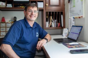 A smiling middle-aged man with glasses and a blue polo shirt sits at an office desk. His left forearm rests on the white desk surface next to an open laptop computer, a computer mouse, and a smartphone. Behind him is a dark wood shelving unit filled with papers, and to his right is a white notice board with printed documents.