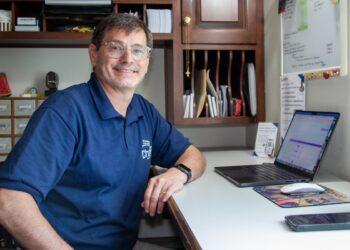 A smiling middle-aged man with glasses and a blue polo shirt sits at an office desk. His left forearm rests on the white desk surface next to an open laptop computer, a computer mouse, and a smartphone. Behind him is a dark wood shelving unit filled with papers, and to his right is a white notice board with printed documents.
