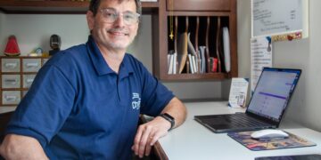 A smiling middle-aged man with glasses and a blue polo shirt sits at an office desk. His left forearm rests on the white desk surface next to an open laptop computer, a computer mouse, and a smartphone. Behind him is a dark wood shelving unit filled with papers, and to his right is a white notice board with printed documents.