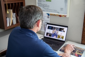 An over-the-shoulder view of a man with graying hair wearing a blue polo shirt, sitting at a desk and looking at an open laptop screen. The laptop shows a grid of several people on a video conference call. His hand rests on a mousepad next to the computer, and a wooden wall organizer and a whiteboard are visible nearby.