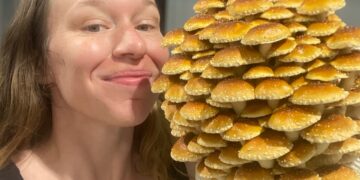 A close-up, indoor shot of a smiling woman standing side-by-side with a massive, dome-shaped cluster of cultivated mushrooms. The woman has long brown hair and is wearing a black tank top. Next to her face, she holds a large fruiting block covered in dozens of overlapping chestnut mushrooms (Pholiota adiposa). The mushrooms feature bright golden-brown, rounded caps speckled with small white textured scales. The background is softly blurred with neutral gray tones.
