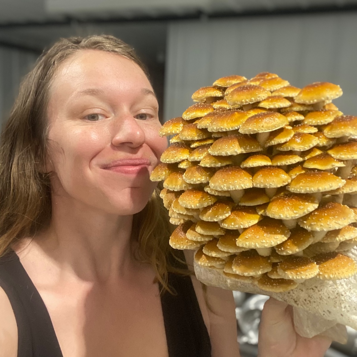 A close-up, indoor shot of a smiling woman standing side-by-side with a massive, dome-shaped cluster of cultivated mushrooms. The woman has long brown hair and is wearing a black tank top. Next to her face, she holds a large fruiting block covered in dozens of overlapping chestnut mushrooms (Pholiota adiposa). The mushrooms feature bright golden-brown, rounded caps speckled with small white textured scales. The background is softly blurred with neutral gray tones.