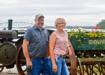 Medium shot of a smiling man and woman standing together in front of vintage farm machinery on a cloudy day. The man on the left has a goatee and wears a gray short-sleeve work uniform shirt with a name patch reading "Ryan," denim jeans, and a camouflage baseball cap. The woman beside him has a blonde bob hairstyle and wears a sleeveless, lace-trimmed pink top and blue jeans, leaning against a large, rusty iron tractor wheel. Behind them is a green wooden vintage "Oliver" implement box repurposed as a planter box filled with vibrant yellow, white, and pink flowers. An agricultural irrigation system structure is visible in the background under an overcast sky.