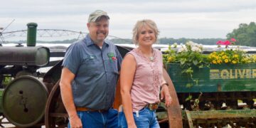 Medium shot of a smiling man and woman standing together in front of vintage farm machinery on a cloudy day. The man on the left has a goatee and wears a gray short-sleeve work uniform shirt with a name patch reading "Ryan," denim jeans, and a camouflage baseball cap. The woman beside him has a blonde bob hairstyle and wears a sleeveless, lace-trimmed pink top and blue jeans, leaning against a large, rusty iron tractor wheel. Behind them is a green wooden vintage "Oliver" implement box repurposed as a planter box filled with vibrant yellow, white, and pink flowers. An agricultural irrigation system structure is visible in the background under an overcast sky.