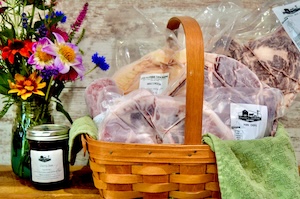 Close-up shot of a farm store gift bundle displayed on a warm wooden tabletop. A shallow woven wooden basket with a high handle holds several clear, vacuum-sealed packages of cuts of meat, lined with a green cloth towel. To the left, a glass mason jar filled with a dark jam or honey sits in front of a green glass jar vase packed with vibrant wildflowers, including red, purple, orange, and blue blooms. The background features a rustic, light-colored wood panel wall.