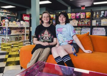 Two smiling young women sit next to each other on a bright orange plush sofa inside a colorful toy and collectible shop. The woman on the left wears glasses and a black t-shirt with a complex graphic design. The woman on the right has dark hair with bangs and wears a light blue Hatsune Miku graphic t-shirt, dark shorts, and striped knee-high socks. The background is filled with retail shelves stocked with various toys, blind boxes, a Hello Kitty plush, and snacks. A black-and-white checkered floor pattern is visible on the left side of the room.