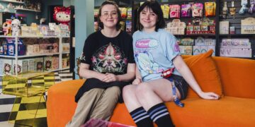 Two smiling young women sit next to each other on a bright orange plush sofa inside a colorful toy and collectible shop. The woman on the left wears glasses and a black t-shirt with a complex graphic design. The woman on the right has dark hair with bangs and wears a light blue Hatsune Miku graphic t-shirt, dark shorts, and striped knee-high socks. The background is filled with retail shelves stocked with various toys, blind boxes, a Hello Kitty plush, and snacks. A black-and-white checkered floor pattern is visible on the left side of the room.