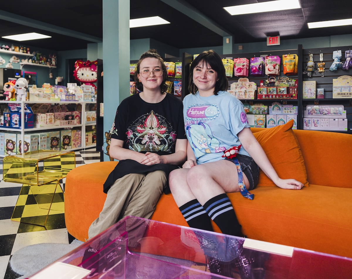 Two smiling young women sit next to each other on a bright orange plush sofa inside a colorful toy and collectible shop. The woman on the left wears glasses and a black t-shirt with a complex graphic design. The woman on the right has dark hair with bangs and wears a light blue Hatsune Miku graphic t-shirt, dark shorts, and striped knee-high socks. The background is filled with retail shelves stocked with various toys, blind boxes, a Hello Kitty plush, and snacks. A black-and-white checkered floor pattern is visible on the left side of the room.
