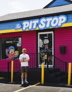 A man stands outside a bright storefront with hot pink siding and a bold blue roof trim. The large, rounded white sign above the door reads "The Pit Stop." The man is smiling, wearing a white t-shirt, dark shorts, white sneakers, and a tan baseball cap with his arms crossed. Black metal railings flank the steps leading up to the main glass entrance door, which features the building number 3840. A large window to the left displays a cartoon character and the word "SNACKS."