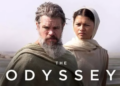 A movie-style promotional image titled "THE ODYSSEY" featuring actors Matt Damon and Zendaya in historical, desert-style clothing. Matt Damon stands in the foreground with a long, graying beard and a draped green cloak. Zendaya stands slightly behind him to the right, looking forward with a serious expression while wearing a cream-colored garment and head wrap. The background is a vast, hazy desert landscape.