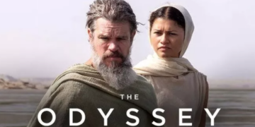 A movie-style promotional image titled "THE ODYSSEY" featuring actors Matt Damon and Zendaya in historical, desert-style clothing. Matt Damon stands in the foreground with a long, graying beard and a draped green cloak. Zendaya stands slightly behind him to the right, looking forward with a serious expression while wearing a cream-colored garment and head wrap. The background is a vast, hazy desert landscape.
