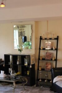 The interior of a dance studio lobby or waiting area featuring a mirror on the wall that reads, "life is better when you DANCE." Below the mirror sits a black cubby shelving unit decorated with a patterned runner and a small vase of white flowers. To the right, a black wooden leaning ladder shelf displays framed photos, golden cups, and candle holders.
