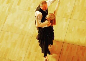 An overhead, bird's-eye view of a ballroom dance couple performing on a light wooden dance floor. The male dancer is wearing a dark vest over a white long-sleeved shirt. The female dancer, with her back mostly to the camera, wears a black dress with subtle embellishments and high heels, her blonde hair styled up as she arches slightly backward in a dance frame.