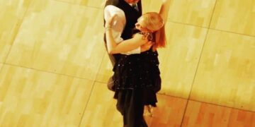 An overhead, bird's-eye view of a ballroom dance couple performing on a light wooden dance floor. The male dancer is wearing a dark vest over a white long-sleeved shirt. The female dancer, with her back mostly to the camera, wears a black dress with subtle embellishments and high heels, her blonde hair styled up as she arches slightly backward in a dance frame.
