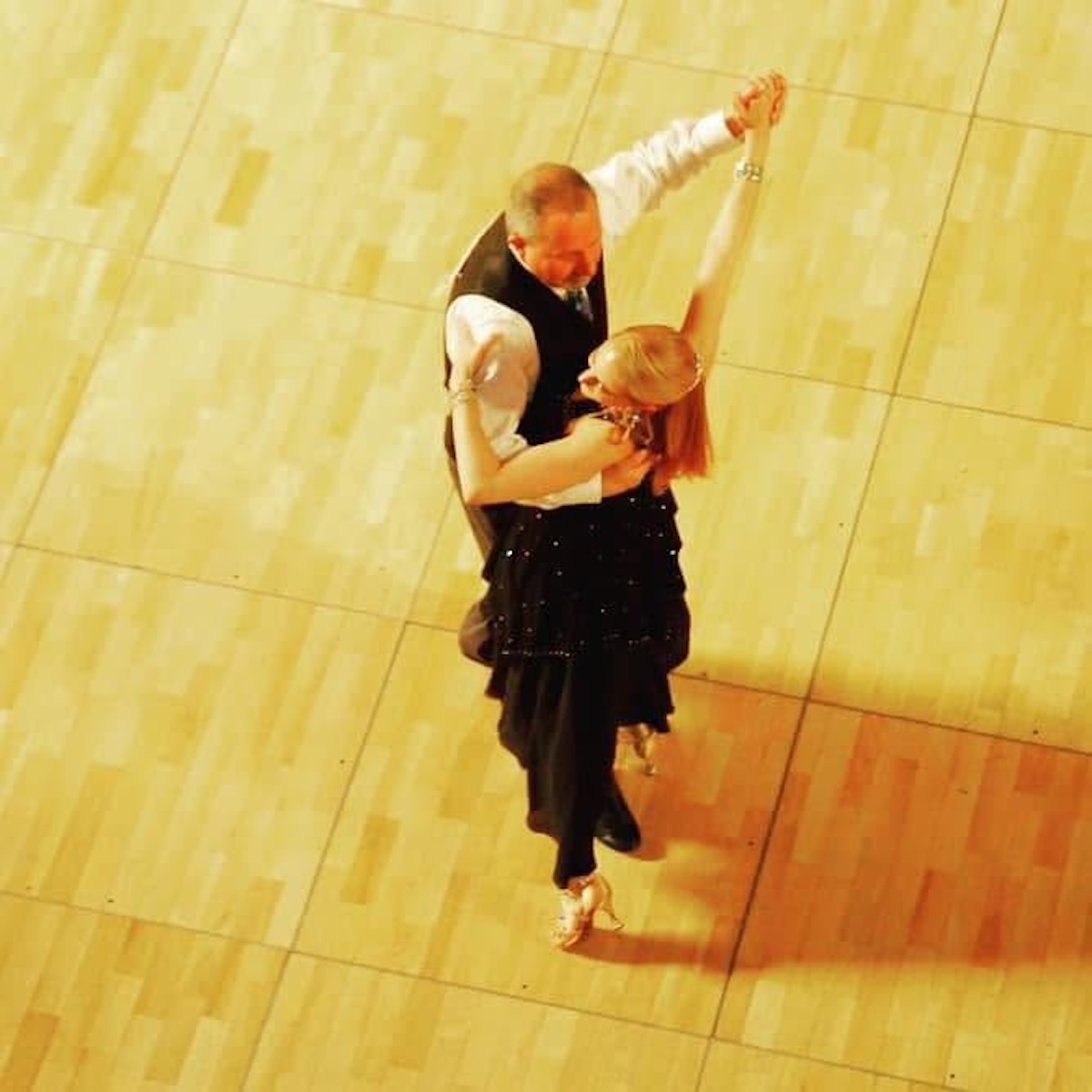 An overhead, bird's-eye view of a ballroom dance couple performing on a light wooden dance floor. The male dancer is wearing a dark vest over a white long-sleeved shirt. The female dancer, with her back mostly to the camera, wears a black dress with subtle embellishments and high heels, her blonde hair styled up as she arches slightly backward in a dance frame.