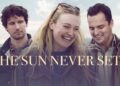 A promotional still and title card for the film "The Sun Never Sets." Dakota Fanning stands in the center, smiling broadly and laughing, wearing a dark green canvas jacket with sunglasses on her head. To her left, Cory Michael Smith looks toward her with a thoughtful expression. To her right, Jake Johnson looks down with a warm smile. They are dressed in casual layers against an outdoor backdrop of pale blue skies and distant mountains. Large, elegant gold text across the center reads "THE SUN NEVER SETS."