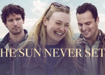 A promotional still and title card for the film "The Sun Never Sets." Dakota Fanning stands in the center, smiling broadly and laughing, wearing a dark green canvas jacket with sunglasses on her head. To her left, Cory Michael Smith looks toward her with a thoughtful expression. To her right, Jake Johnson looks down with a warm smile. They are dressed in casual layers against an outdoor backdrop of pale blue skies and distant mountains. Large, elegant gold text across the center reads "THE SUN NEVER SETS."