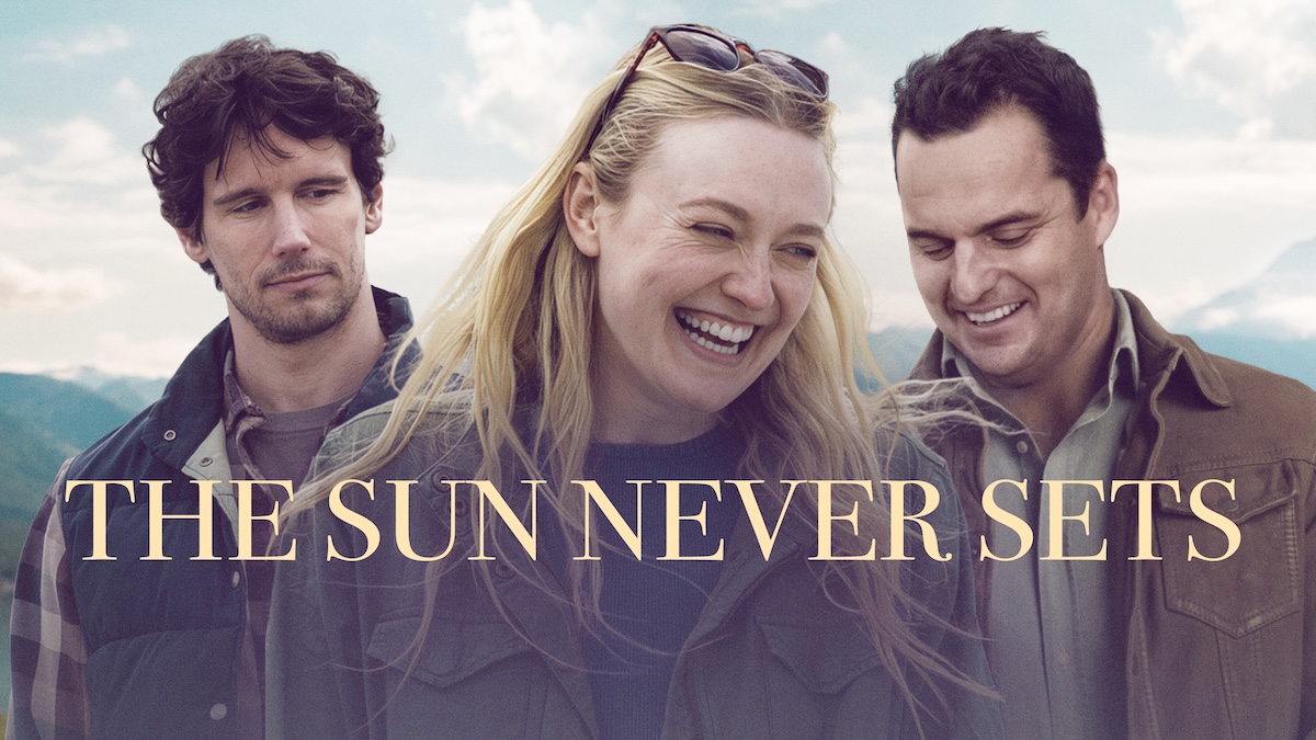 A promotional still and title card for the film "The Sun Never Sets." Dakota Fanning stands in the center, smiling broadly and laughing, wearing a dark green canvas jacket with sunglasses on her head. To her left, Cory Michael Smith looks toward her with a thoughtful expression. To her right, Jake Johnson looks down with a warm smile. They are dressed in casual layers against an outdoor backdrop of pale blue skies and distant mountains. Large, elegant gold text across the center reads "THE SUN NEVER SETS."