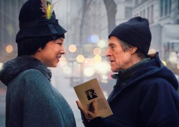 A movie poster for "Late Fame" featuring actors Willem Dafoe and Greta Lee standing face-to-face on a city street. The two characters smile warmly at each other amidst a soft-focus urban background with blurred lights. Greta Lee is on the left, wearing a dark coat and a black vintage-style hat adorned with a colorful feather. Willem Dafoe is on the right, dressed in a dark blue hooded coat and a beanie, holding a small book or pamphlet between them. The film's title, "LATE FAME," is written in large gold letters at the bottom, with the actors' names printed above it.