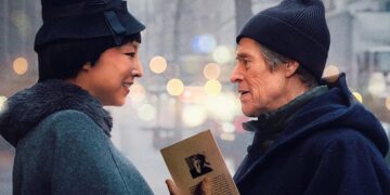 A movie poster for "Late Fame" featuring actors Willem Dafoe and Greta Lee standing face-to-face on a city street. The two characters smile warmly at each other amidst a soft-focus urban background with blurred lights. Greta Lee is on the left, wearing a dark coat and a black vintage-style hat adorned with a colorful feather. Willem Dafoe is on the right, dressed in a dark blue hooded coat and a beanie, holding a small book or pamphlet between them. The film's title, "LATE FAME," is written in large gold letters at the bottom, with the actors' names printed above it.