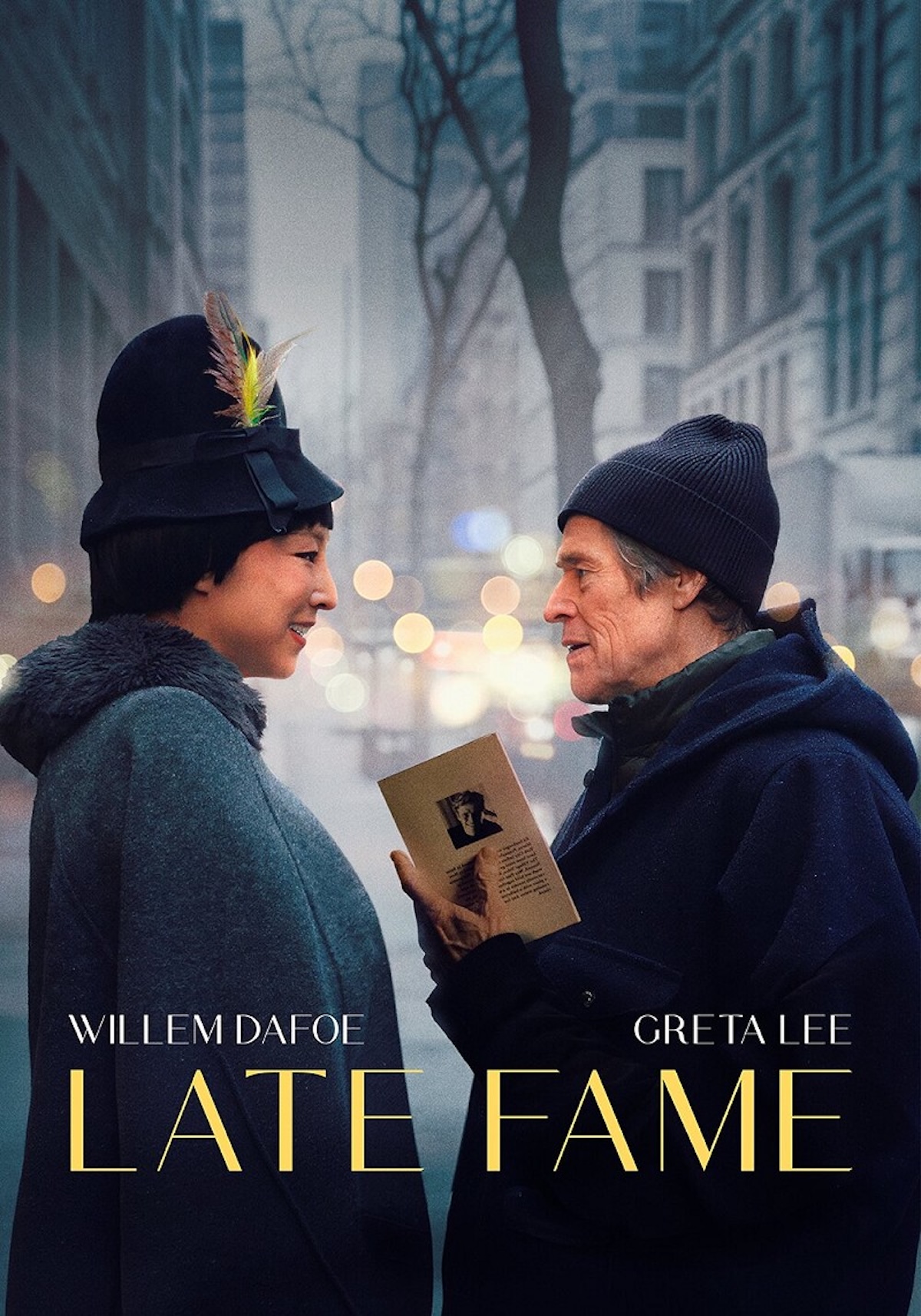 A movie poster for "Late Fame" featuring actors Willem Dafoe and Greta Lee standing face-to-face on a city street. The two characters smile warmly at each other amidst a soft-focus urban background with blurred lights. Greta Lee is on the left, wearing a dark coat and a black vintage-style hat adorned with a colorful feather. Willem Dafoe is on the right, dressed in a dark blue hooded coat and a beanie, holding a small book or pamphlet between them. The film's title, "LATE FAME," is written in large gold letters at the bottom, with the actors' names printed above it.