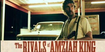 A close-up shot of actor Matthew McConaughey in the film "The Rivals of Amziah King." He stands under a brightly lit carport at night, looking off-camera with a serious expression. He has longer curly hair, a beard, and wears a blue bandana, a light yellow t-shirt, and dark suspenders with a rifle strapped to his chest. A vintage white pickup truck sits in the background under the fluorescent lights.